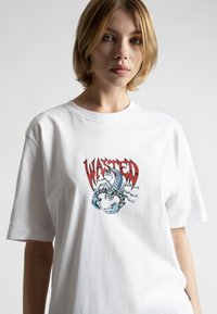 Bílý bavlněný tričko s grafickým potiskem modrého škorpiona a červeného textu "WASTED", krátké rukávy a volný střih.