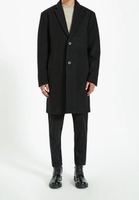 Cappotto in lana nera con rever a punta, due bottoni e tasche laterali, abbinato a pantaloni neri e stivali in pelle. Texture liscia, vestibilità su misura.