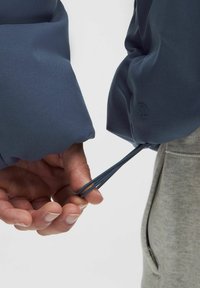 Modrá športová bunda s elastickými manžetami, prezentujúca textúrovanú tkaninu a šnúrky, držená v ruke oproti šedej bunde.