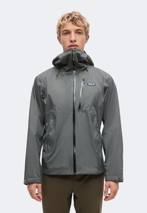 Junger Mann trägt eine graue wasserdichte Jacke mit Kapuze, die bis zum Kinn zugezogen ist, und steht vor einem einfarbigen hellen Hintergrund.