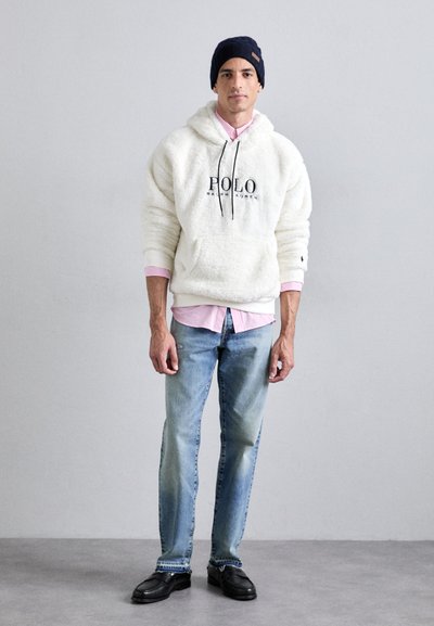 Hoodie de felpa branca com logotipo preto, sobre uma camisa às riscas rosa. Calças de ganga azul-claro e sapatos pretos completam o visual, rematado por um gorro azul-marinho.