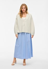 Šviesiai pilkas, per didelis megztinis su V formos iškirpte ir plačiomis rankovėmis, derinamas su ilga, šviesiai mėlyna sijonu iš plazdančio audinio. Šviesaus atspalvio šlepetės.