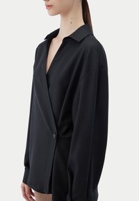 Svart blus med lång ärm, V-hals med slag, omlottdesign och en enkel knappstängning. Tillverkad av mjukt tyg med avslappnad passform.