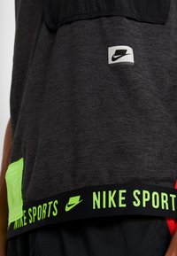 Sportowy top Nike w ciemnoszarym kolorze, z czarną siateczką u góry, neonowozielonymi akcentami oraz elastycznym paskiem z logo "NIKE SPORTS".