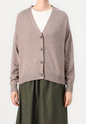 Personne portant un cardigan boutonné col en V couleur taupe par-dessus un haut blanc et une jupe vert olive, debout devant un fond uni.