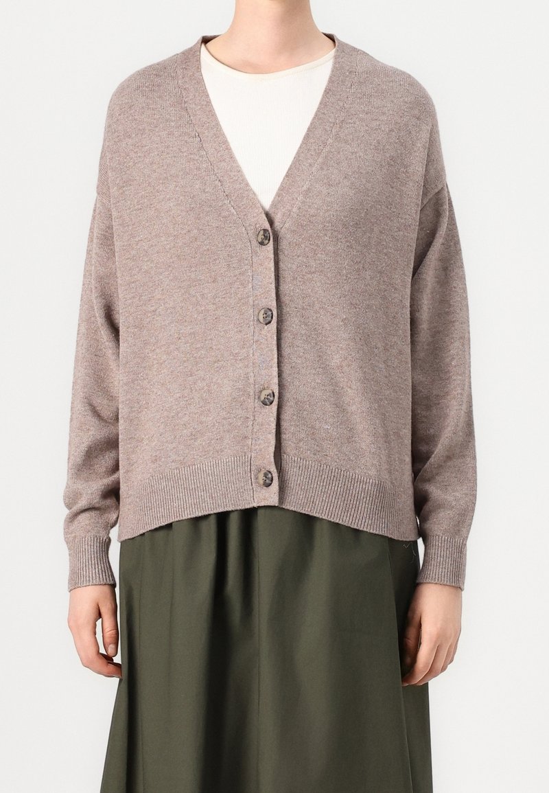 Personne portant un cardigan boutonné col en V couleur taupe par-dessus un haut blanc et une jupe vert olive, debout devant un fond uni.