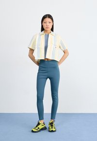 Gul og hvit stripete skjorte som er avkortet over en blå tank, kombinert med teal leggings og svarte og gule joggesko. Enkel design, rene linjer.
