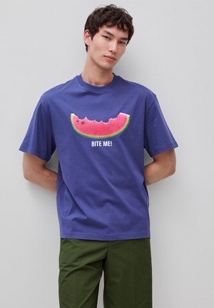 Junger Mann trägt ein blaues T-Shirt mit einer angebissenen Wassermelonen-Grafik und dem Text „BITE ME!“, kombiniert mit grünen Hosen, steht vor einer grauen Wand.