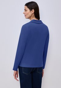 Femme aux longs cheveux foncés portant une chemise bleue à manches longues et un jean foncé, debout de profil à gauche sur un fond clair uni.