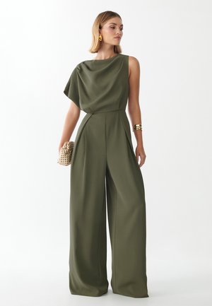 Frau trägt einen olivgrünen One-Shoulder-Jumpsuit mit weitem Bein, hält eine geflochtene Clutch und trägt goldenen Schmuck vor weißem Hintergrund.