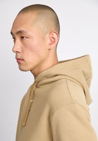 Sweatshirt bege com capuz, feito de um tecido texturizado, apresentando um cordão dourado e um pequeno logo branco na frente. Ajuste suave e casual.