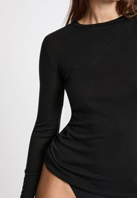 Primo piano di una persona che indossa una maglietta nera aderente a maniche lunghe e pantaloni neri su uno sfondo chiaro e uniforme.