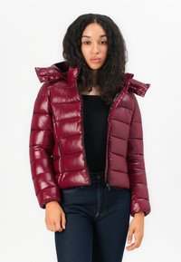 Bordeaux pufferjack met glanzende afwerking, horizontaal gebiesde patroon, rits aan de voorkant en afneembare capuchon. Gecombineerd met een zwarte top en donkere jeans.