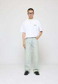 T-shirt bianco oversize con piccolo logo nero, jeans azzurri a gamba larga, scarpe nere e grandi occhiali con montatura viola.