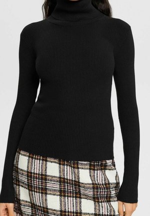 Kobieta w dopasowanym czarnym prążkowanym golfie i wysokim stanem białej, czarnej i musztardowej spódnicy w kratę.