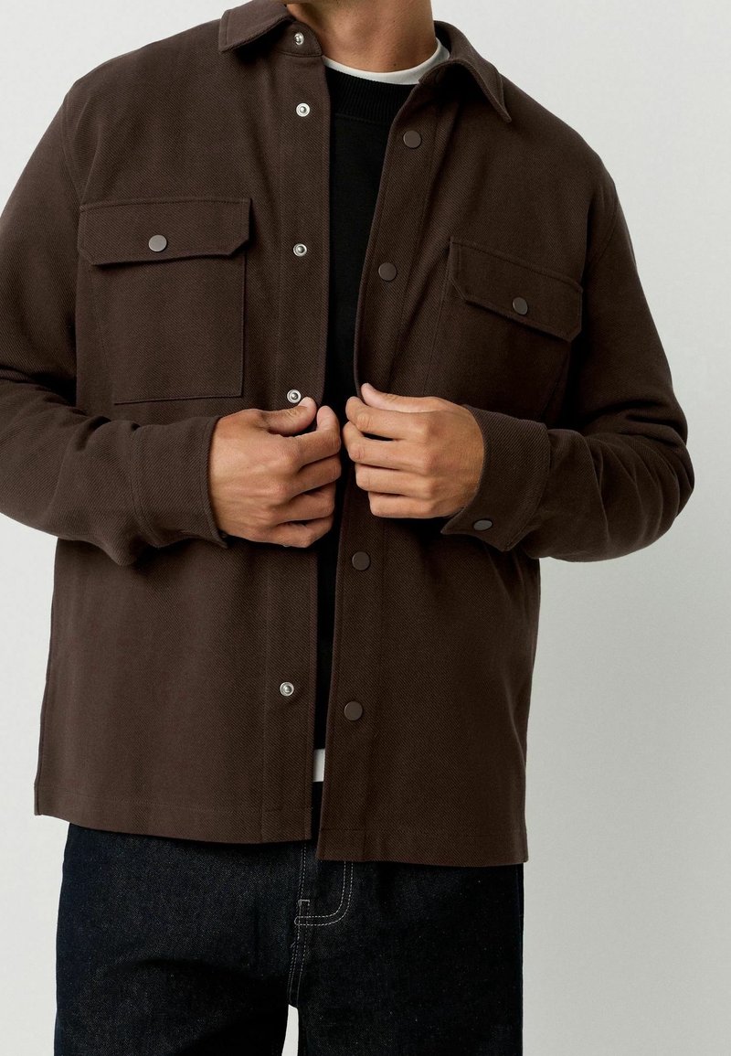 Homme portant une veste marron foncé boutonnée avec des poches à rabat, boutonnant les boutons sur une chemise noire et un jean foncé.