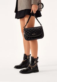 Bolso de mano negro acolchado con un logo dorado, cierre de cremallera y correa corta, combinado con botas de tobillo negras brillantes que presentan acentos de cadena dorada.