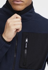 Flis jakna u tamnoplavoj boji s crnim zippered džepom na prsima. Ima visoki ovratnik i elastične manžete. Mekana tekstura i kroj koji se prilagođava.