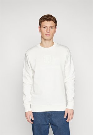 Hvid sweatshirt lavet af glat stof, med en ribbet rund hals og manchetter, samt et præget logo på fronten. Kombineret med mørke jeans.