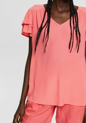Personne à la peau foncée avec de longues tresses portant un chemisier rose corail à manches volantées et un pantalon assorti.