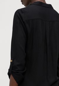 Chemise noire à manches longues avec un col classique, des manches retroussées avec détail de bouton et un pli au dos pour plus de confort, le tout dans un tissu léger.