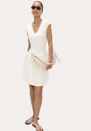 Vrouw draagt een witte mouwloze jurk met V-hals, bruine zonnebril, draagt een beige schoudertas en draagt beige slip-on sandalen met uitsnijdingen.