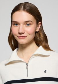 Cream quarter-zip pullover sweater with navy stripes, made of soft fabric. Features a silver zipper and a small logo on the chest.