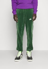 Grønne fløjlstrækbukser med hvide side-striber og Nike-logo, parret med sort-hvide høj-top sneakers med grønne accenter.