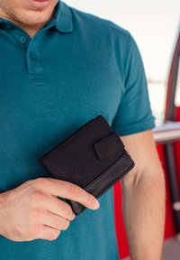 Black wallet made of fabric and leather with a clasp. It features a minimalistic design, held in a hand against a teal shirt.