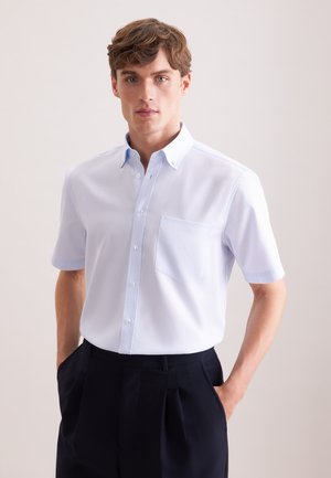 Jonge man staand met handen in zijn zakken, gekleed in een lichtblauw kortermig overhemd met knopen en een donkere broek, tegen een effen achtergrond.