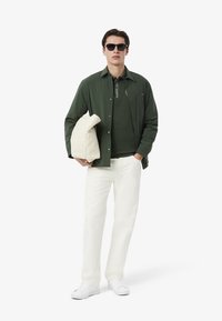 Zielona kurtka zapinana na guziki noszona na ciemnozielonym polo, białe spodnie i białe sneakersy; trzyma beżową torbę. Dodatki to ciemne okulary przeciwsłoneczne.