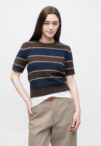 Mladi žena s dugom smeđom kosom nosi kratku majicu s prugama od mekanog materijala, bež hlače i smeđi remen, pozirajući ispred bijele pozadine.