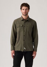 Olive green long-sleeve shirt with two chest pockets, button front, and textured fabric. Paired with black pants.