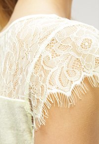 Krajkový top s propracovanými květinovými vzory, barvy slonové kosti, s třásněmi na okrajích. Tkanina je překryta měkkým, světle zeleným základem.