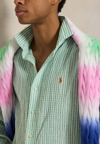 Férfi zöld-fehér csíkos ingben, narancssárga logóval, és egy rózsaszín-fehér-kék átmenetes kötött pulóver vállra dobva.