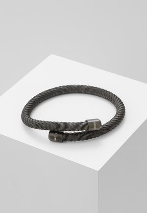 Black flexible bracelet with a coiled design, featuring cylindrical metal ends with engraved branding, displayed on a minimal white pedestal.