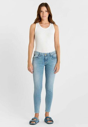 Femme debout portant un haut blanc sans manches, un jean skinny bleu clair et des sandales bleues à bouts ouverts sur un fond clair uni.