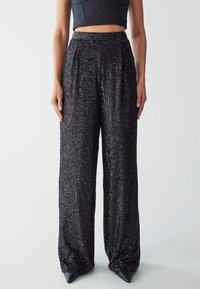 Pantaloni a gamba larga neri in paillettes con una superficie testurizzata e scintillante, dotati di vita alta e pieghe, abbinati a scarpe nere appuntite.