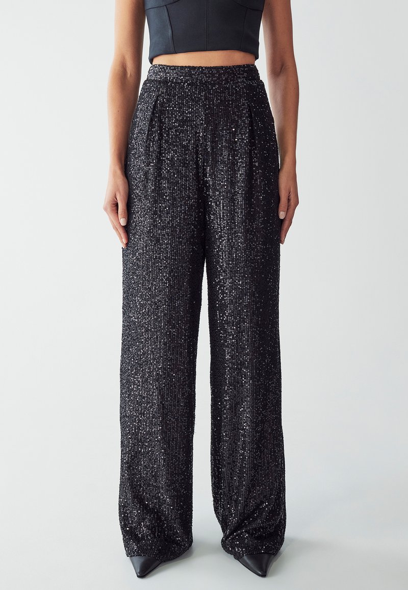 Pantaloni a gamba larga neri in paillettes con una superficie testurizzata e scintillante, dotati di vita alta e pieghe, abbinati a scarpe nere appuntite.