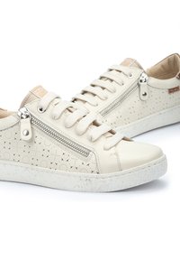 Zapatillas beige con diseño perforado, acentos de cremallera plateada, cordones planos y suela blanca texturizada. Cuero suave con un ligero brillo.