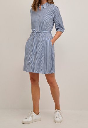 Vrouw draagt een blauw-wit verticaal gestreepte blousejurk met ceintuur in de taille, driekwartmouwen en witte sneakers. Handen in de zakken.