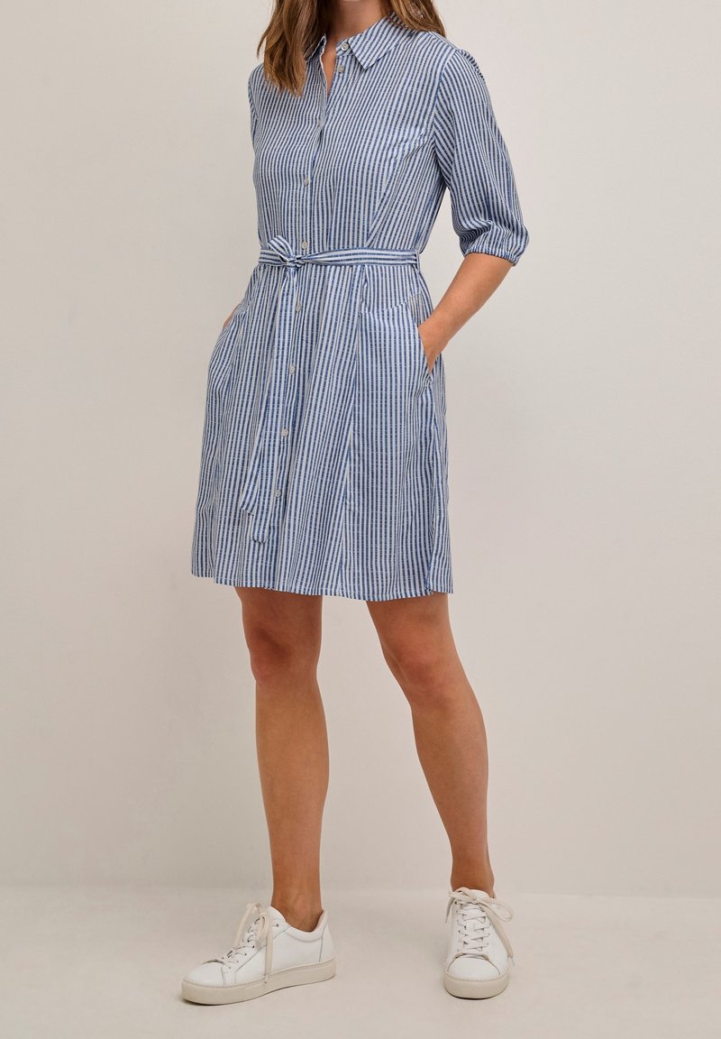 Femme portant une robe chemise à rayures verticales bleu et blanc avec ceinture à la taille, manches trois-quarts et baskets blanches. Mains dans les poches.