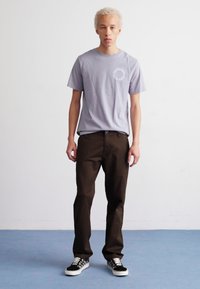 Graues T-Shirt mit kurzen Ärmeln und Logo, braune gerade Hosen und schwarz-weiße kariertes Sneakers. Das Model steht auf einem blauen Boden.