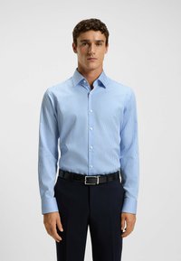 Camicia a maniche lunghe a righe blu chiaro con colletto, chiusura a bottoni e tessuto morbido. Indossata con pantaloni scuri e una cintura nera.