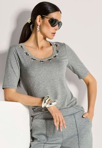 Frau mit dunklem Pferdeschwanz, die schwarze Sonnenbrille, ein grau verziertes Oberteil, passende Hose, ein großes silbernes Armband und hängende Ohrringe trägt, posiert im Lean.