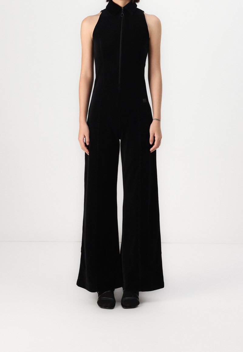 Jumpsuit en velours noir avec col montant, devant zippé et jambes larges. Présente un design élégant et une texture lisse. Aucune motif ni accent visible.