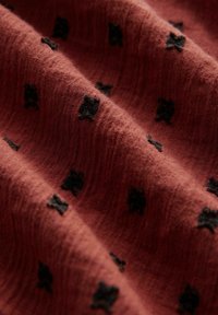 Tissu couleur rouille à la texture douce, présentant des motifs floraux noirs. Le matériau semble léger et légèrement froissé.