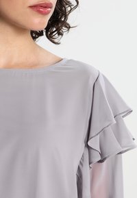 Blusa grigia chiaro con scollatura tonda e maniche a volant stratificate, realizzata in un tessuto liscio con una consistenza morbida.
