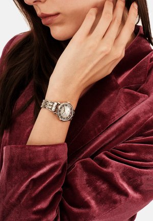 Sølvur med rund urskive, rustfri stålkæde og grå urskive. Båret på et håndled imod et burgunderfarvet velourbeklædning.