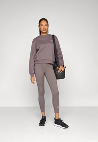 Sieviete stāv, tērpusies mauve krāsas džemperī un legingos, ar melnām sporta kurpēm un melnu pleca somu, pret vienkāršu gaiši pelēku fonu.